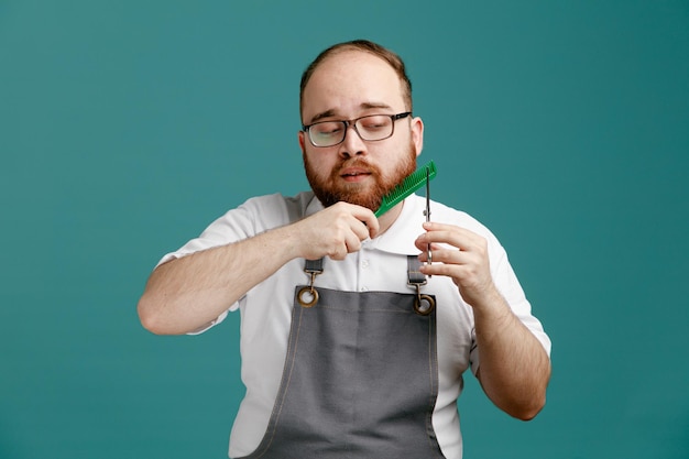 Concentrado jovem barbeiro vestindo uniforme e óculos penteando a barba com pente teaser segurando e olhando para tesoura isolada em fundo azul