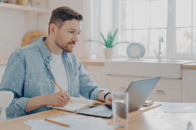 Concentrado jovem empresário escrevendo notas na agenda e trabalhando remotamente no laptop na internet de casa enquanto está sentado à mesa da cozinha. Conceito de freelance e trabalho online