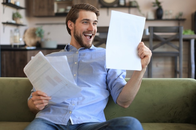 Concentrado jovem empresário freelancer sentado no sofá com o laptop e examinando documentos.