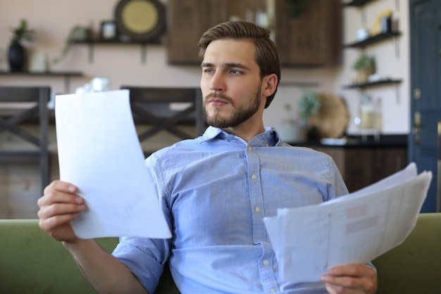 Concentrado jovem empresário freelancer sentado no sofá com o laptop e examinando documentos.