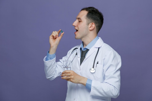 Concentrado jovem médico masculino vestindo roupão médico e estetoscópio no pescoço em pé na vista de perfil segurando cápsula e copo de água em outra mão se preparando para tomar pílula