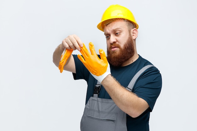 Concentrado jovem trabalhador da construção civil usando capacete de segurança e uniforme tirando as luvas de segurança olhando para a mão isolada no fundo branco