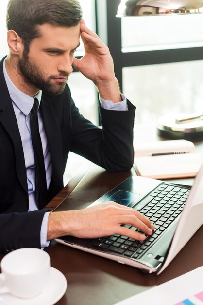 Concentrado no trabalho. Jovem concentrado em um terno tocando a cabeça com a mão e olhando para o laptop enquanto está sentado em seu local de trabalho