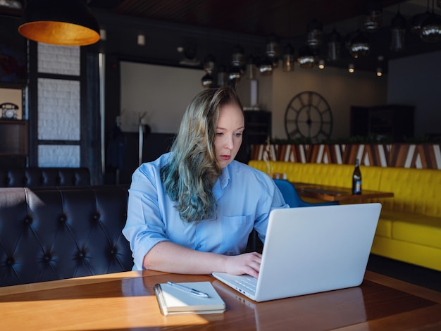 Concentrado no trabalho. Mulher jovem confiante e tamanho em roupas casuais elegantes, trabalhando em um laptop enquanto está sentado perto de uma janela em um café criativo