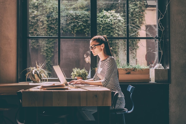 Concentrado no trabalho. Mulher jovem confiante em roupas casuais inteligentes, trabalhando em um laptop, enquanto está sentado perto de uma janela em um escritório criativo ou um café