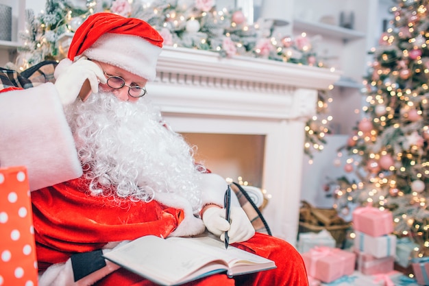 Concentrado y reflexivo, Santa Claus se sienta en la chimenea y el árbol de Navidad escribiendo en un cuaderno