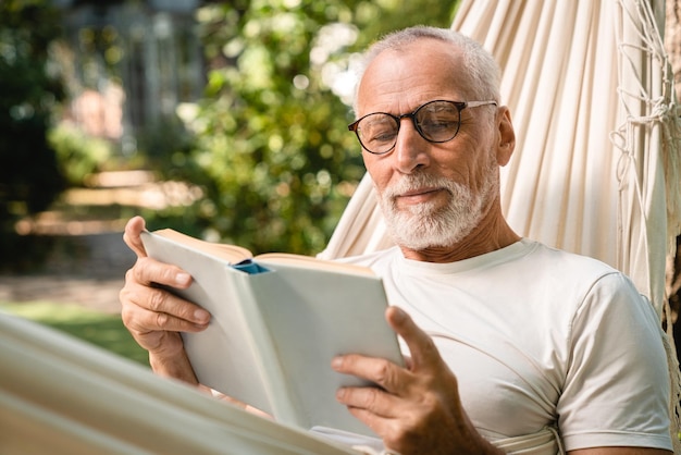 Concentrado soñador viejo anciano anciano abuelo caucásico hombre relajándose descansando en una hamaca mientras lee un libro al aire libre en el bosque del jardín del parque