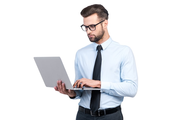 Concentrado en el trabajo. Hombre guapo joven confidente en camisa y corbata trabajando en la computadora portátil mientras está de pie contra el fondo blanco