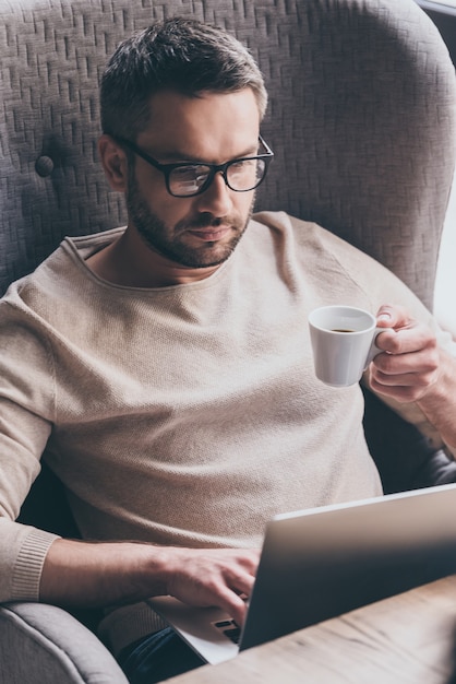 Concentrado en el trabajo. Hombre guapo usando su computadora portátil y sosteniendo la taza de café mientras está sentado en una silla