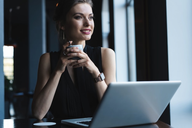 Concentrado en el trabajo. Mujer joven confiada en ropa de sport elegante que trabaja en la computadora portátil mientras que se sienta cerca de la ventana en la oficina creativa o el café.