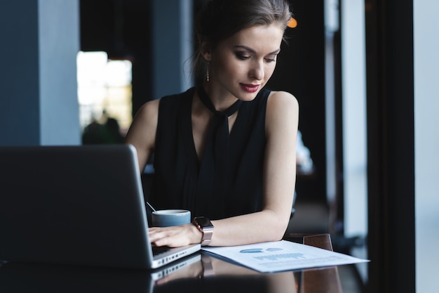 Concentrado en el trabajo. Mujer joven confiada en ropa de sport elegante que trabaja en la computadora portátil mientras que se sienta cerca de la ventana en la oficina creativa o el café.