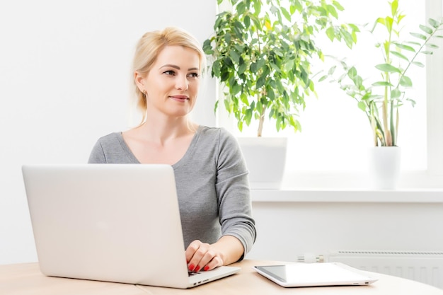 Concentrado en el trabajo. Mujer joven segura de sí misma con ropa informal inteligente que trabaja en una laptop mientras se sienta cerca de la ventana.