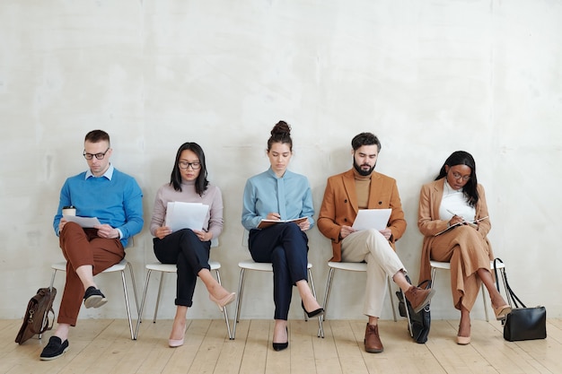 Foto concentrados jovens candidatos multiétnicos para empregos sentados com papéis no corredor e examinando-os antes da entrevista