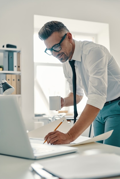 Concentrarse en el trabajo. Apuesto joven en camisa y corbata escribiendo algo y sonriendo mientras está de pie en la oficina