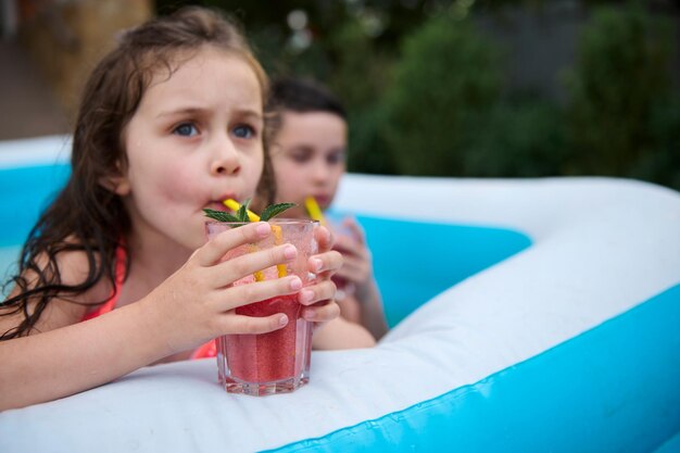 Concentre-se em um copo com suco de frutas frescas nas mãos de uma menina desfocada bebendo coquetel em uma piscina inflável