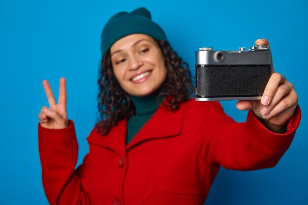 Concentre-se na câmera vintage de equipamento fotográfico de estilo retro nas mãos de uma mulher bonita turva, mostrando o sinal de paz e sorrindo com um sorriso cheio de dentes enquanto toma selfie, contra o fundo de cor azul.
