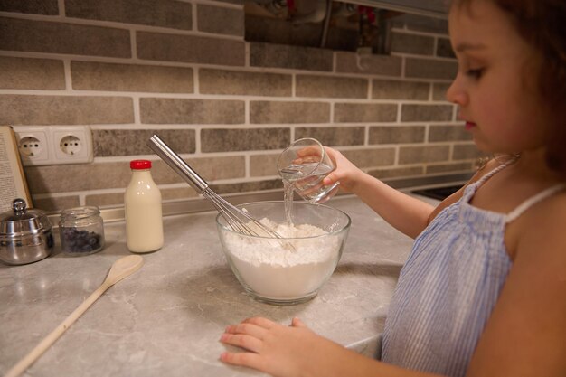 Concentre-se na mão da adorável garotinha, segurando um copo com água e despejando-o em uma tigela com farinha para preparar massa líquida para panquecas shrove na cozinha de casa.