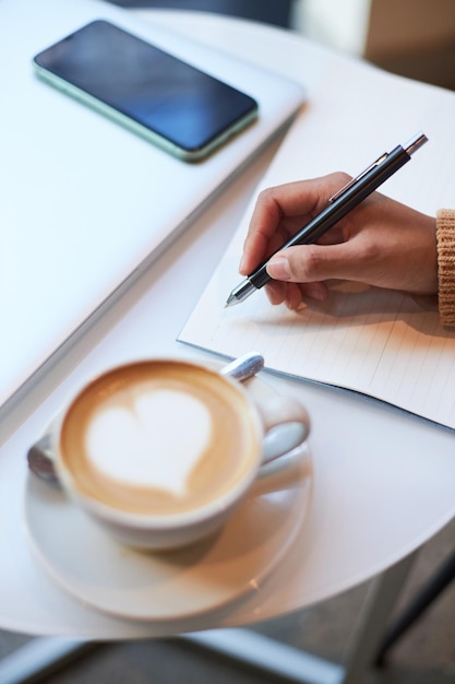 Concentre-se na mão feminina segurando uma caneta de tinta e escrevendo em uma folha de papel branco durante uma pausa para o café