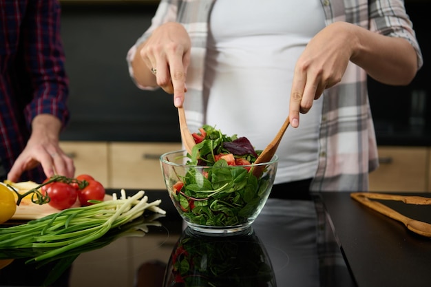 Concentre-se nas mãos da mulher grávida segurando colheres de pau e misturando ingredientes em uma tigela de vidro, preparando uma deliciosa salada saudável para o jantar na ilha da cozinha ao lado do marido cortando legumes