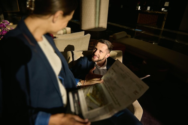 Concéntrese en un apuesto hombre de negocios caucásico mirando a su borrosa socia de negocios, sentado frente a él y leyendo el periódico en el vestíbulo de un hotel