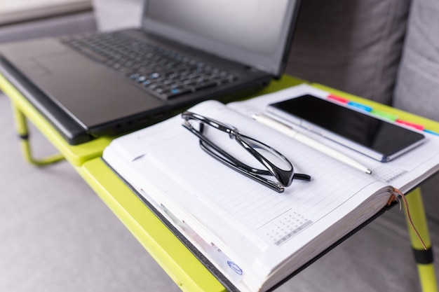 Concéntrese en las gafas que se encuentran en la mesa de la computadora portátil cerca de la página en blanco abierta de un diario con un bolígrafo para hacer citas, organizar un horario