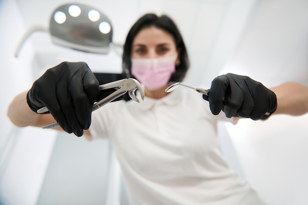 Concéntrese en los instrumentos dentales de acero inoxidable, las pinzas de extracción y el espejo dental en las manos del dentista borroso con guantes quirúrgicos y máscara médica inclinada hacia el paciente para un examen