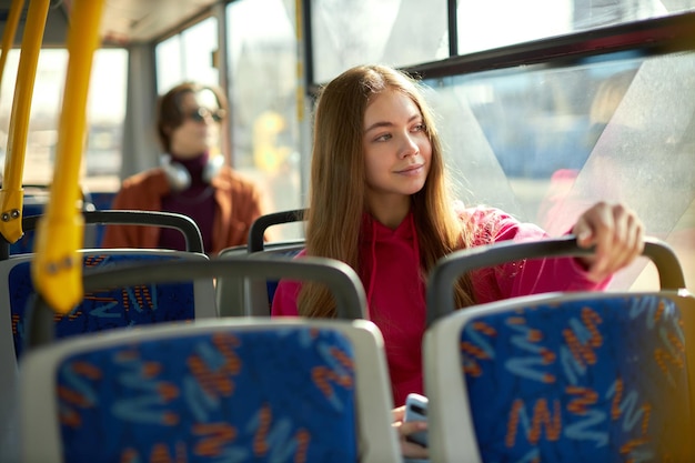 Concéntrese en una joven con capucha rosa sentada en el tranvía con un pasajero borroso en el fondo conveniente