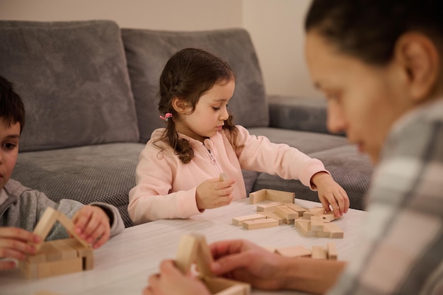 Concéntrese en una linda niña caucásica que construye construcciones de madera con bloques y ladrillos, que desarrolla sus habilidades motoras finas, se sienta a la mesa cerca de su hermano y mamá que se concentra en construir estructuras