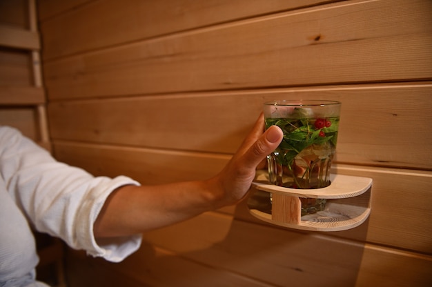 Concéntrese en las manos femeninas sosteniendo un vaso con bebidas vitamínicas mientras se relaja en una acogedora sauna de madera. Agua con menta y bayas en manos de una mujer relajándose en una sauna