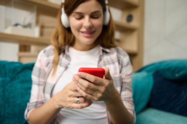 Concéntrese en las manos de una hermosa mujer caucásica borrosa usando un teléfono móvil escuchando música con auriculares inalámbricos mientras se relaja en el sofá de su casa disfrutando de su embarazo