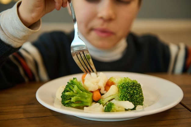 Concéntrese en un plato de verduras al vapor, una comida saludable de brócoli y zanahorias contra un niño borroso comiendo sentado en una mesa y sosteniendo un tenedor. Nutrición, dieta vegetariana para niños pequeños.