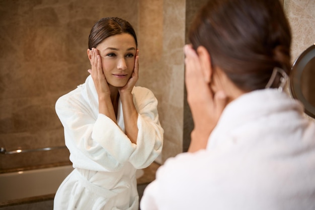 Concéntrese en el reflejo del espejo de la hermosa mujer morena de mediana edad segura con albornoz y tocándose la cara mirándose de pie en el baño durante la rutina de higiene matutina