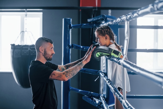 Concepção de assistência. Jovem treinador de boxe tatuado ensina uma linda garotinha no ginásio.