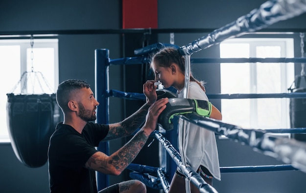 Foto concepción de la asistencia. entrenador de boxeo tatuado joven enseña a niña linda en el gimnasio.