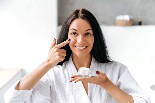 Foto concept de belleza hermosa bien cuidada mujer caucásica morena en una bata de baño blanca con un frasco de crema en la mano aplica una crema hidratante antienvejecimiento en su cara se ocupa de la piel y la juventud