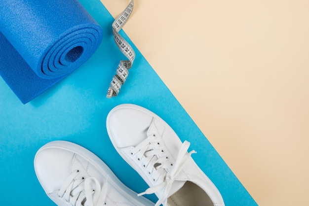 El concepto de accesorios deportivos Una foto de zapatillas blancas, pesas azules, una alfombra de ejercicio azul y otros equipos deportivos contra un fondo beige pastel Vista desde arriba