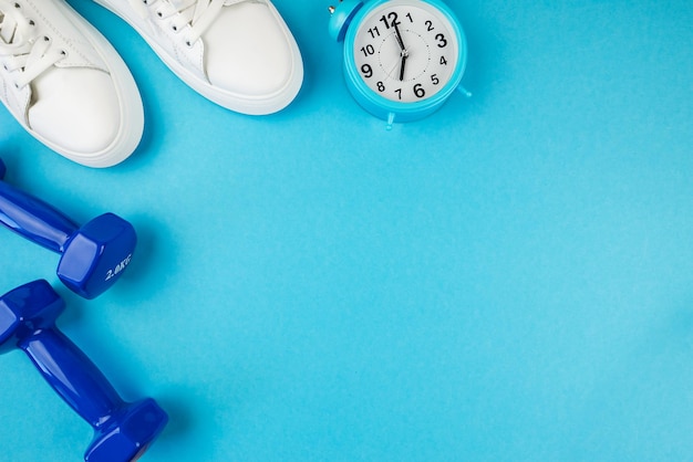 Concepto de accesorios de fitness Foto de vista superior de zapatillas blancas con reloj despertador y mancuernas azules sobre fondo azul aislado con espacio de copia Es hora de entrenar