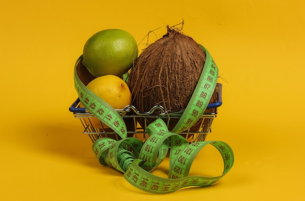 El concepto de adelgazamiento. Frutas tropicales y cinta métrica en la cesta de la compra sobre fondo amarillo. Alimentación saludable. Dieta de frutas. Vista superior