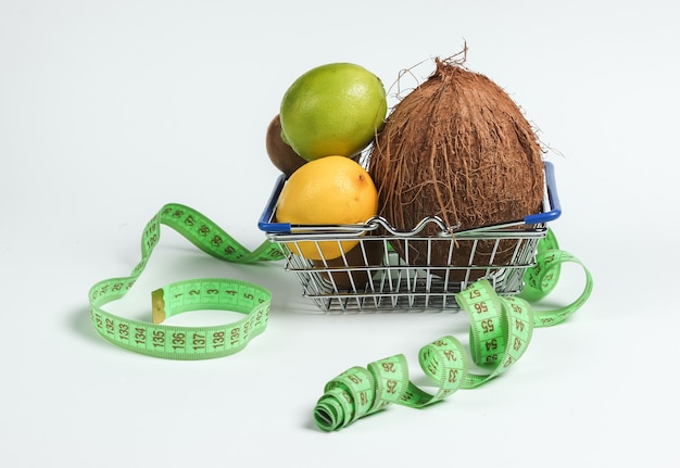 El concepto de adelgazamiento. Frutas tropicales y cinta métrica en la cesta de la compra sobre fondo blanco. Alimentación saludable. Dieta de frutas. Vista superior