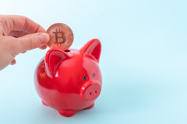 Concepto de ahorro de criptomonedas. Una mano sostiene una moneda bitcoin sobre una hucha roja sobre un fondo azul.