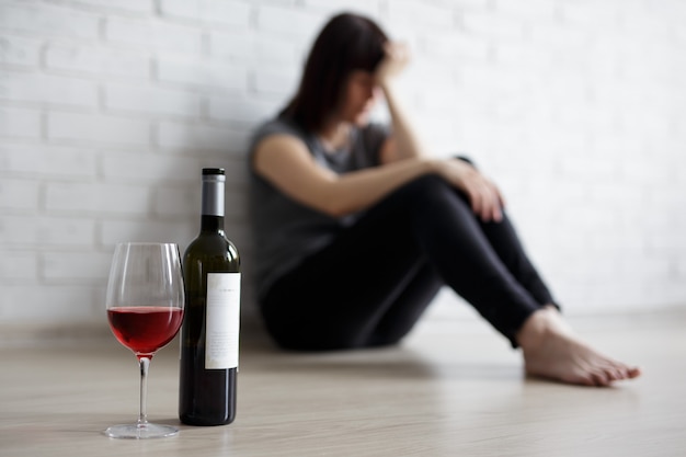 Concepto de alcoholismo femenino, relación, divorcio y depresión - cerca de una copa y una botella de vino y una mujer estresada llorando y sentada en el suelo en casa