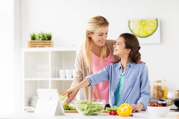 Concepto de alimentación saludable, familia y personas: madre feliz e hija cocinando verduras para la cena usando una receta en línea en una tableta en la cocina de casa