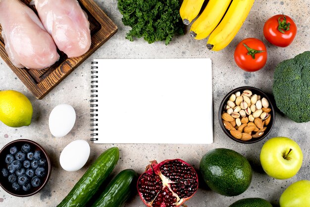 Foto el concepto de alimentos de dieta ceto, filete de pollo, huevos, verduras verdes, tomates, nueces y frutas en un fondo de mesa de hormigón claro, vista superior plana, espacio de copia, maqueta con un cuaderno blanco vacío.