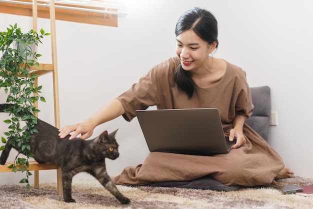 Concepto de amante de las mascotas Mujer asiática joven que usa una computadora portátil y juega con el gato en la sala de estar en casa