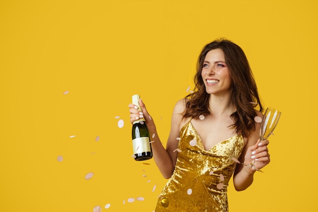 Foto concepto de ambiente festivo mujer feliz vestida con botella de champán y vaso lleno en la mano posando con confeti cayendo