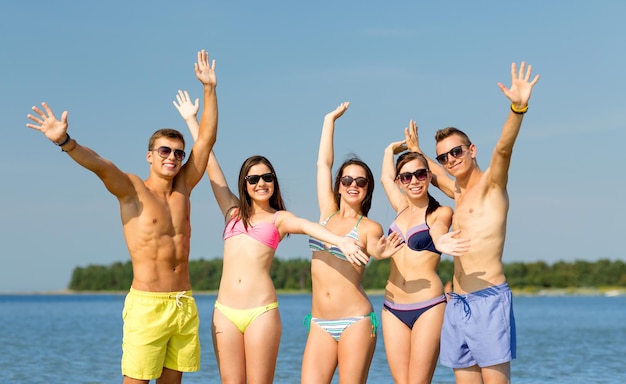 concepto de amistad, mar, vacaciones, gestos y personas - grupo de amigos sonrientes que usan trajes de baño y gafas de sol que agitan las manos en la playa