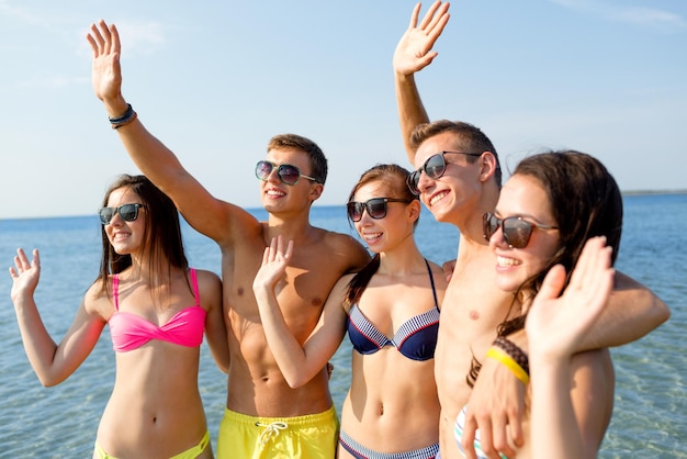 concepto de amistad, mar, vacaciones, gestos y personas - grupo de amigos sonrientes que usan trajes de baño y gafas de sol que agitan las manos en la playa