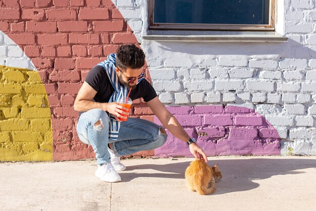 Concepto de amistad y mascotas - Hombre guapo juega con lindo gato rojo al aire libre.