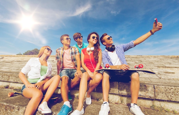 concepto de amistad, ocio, verano, tecnología y personas - grupo de amigos sonrientes con monopatín y smartphone haciendo selfie al aire libre