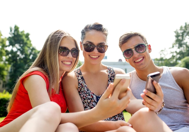 concepto de amistad, ocio, verano, tecnología y personas - grupo de amigos sonrientes con smartphone sentados en el césped y haciendo selfie en el parque